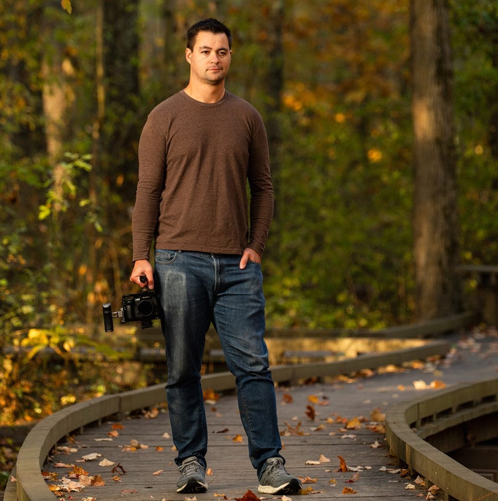 Joe Monzo standing on path holding a camera looking stoic in the woods